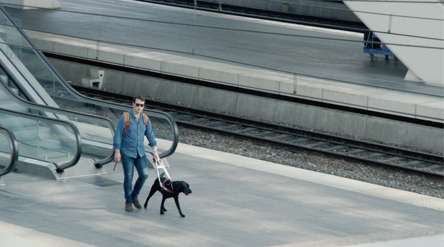 Victor loopt met zijn zwarte blindgeleidehond op het perron van het Luikse treinstation