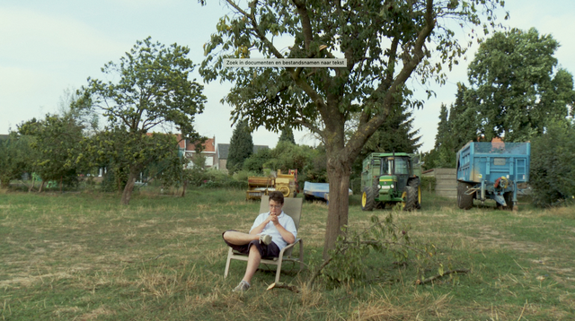 Victor zit in een ligstoel onder een boom in een weide. Achter hem staan verschillende landbouwmachines.