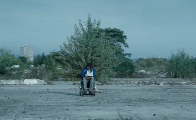Rupaszov, one of the main characters, is in his wheelchair on an abandoned terrain. The terrain is surrounded by trees. Rupaszov is keeping his head down.