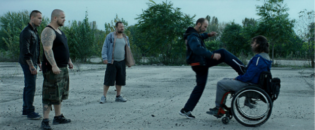 Main character Rupaszov is in his wheelchair. He gets kicked in the stomach by another man but does not react. Behind the attacker are three more men. They are on an abandoned terrain surrounded by trees.