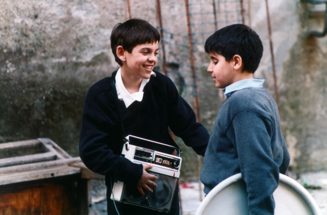 Mirco staat tegenover een andere jongen, die een oude cassetespeler vastheeft. De jongen lacht en steekt zijn hand uit naar Mirco.
