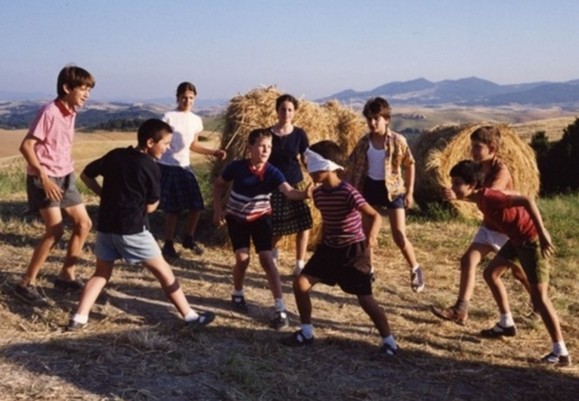 A group of boys and one girl are in a field. They are all standing around Mirco, who is blindfolded. In the background are hay bales and mountains.