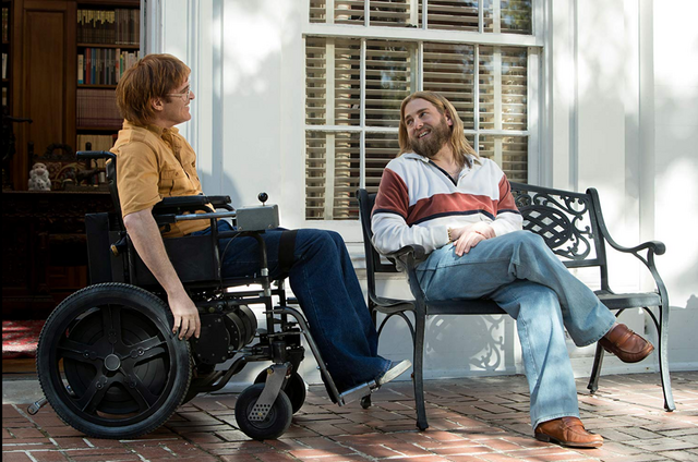 John zit in zijn rolstoel. Naast hem zit een man op een bankje. Ze lachen.