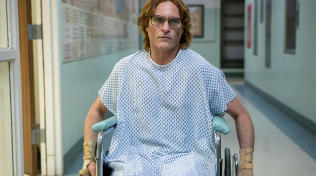 John duwt zichzelf voort door de gang van het ziekenhuis in een rolstoel. Hij draagt een ziekenhuiskleed en kijkt boos.