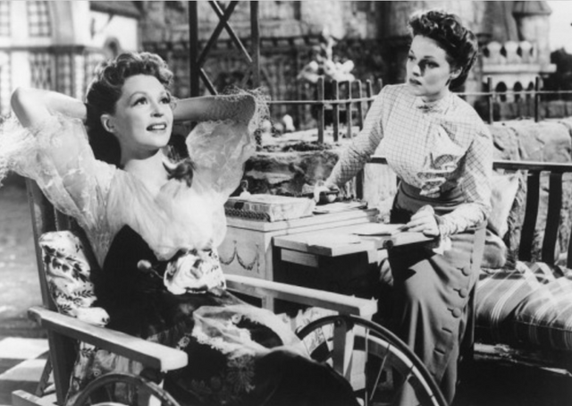 Black and white image of two young women. One of the women is in a wheelchair. She is holding her hands behind her head and is laughing. The other woman is getting up to help her, looking rather concerned.