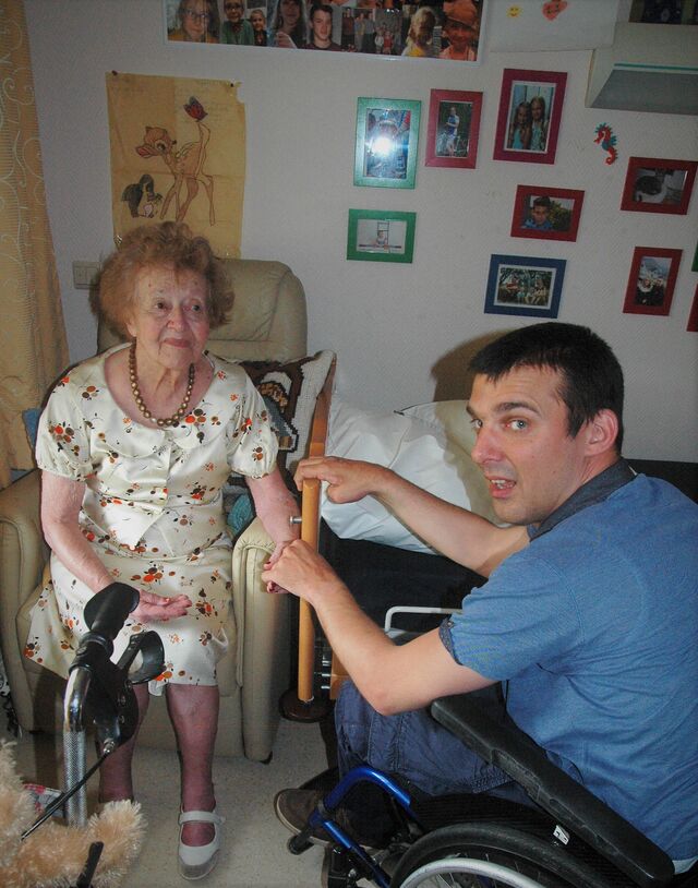 Een bejaarde vrouw zit in een zetel. Schuin tegenover haar zit een jongeman in een rolstoel. Ze houden elkaars hand vast.