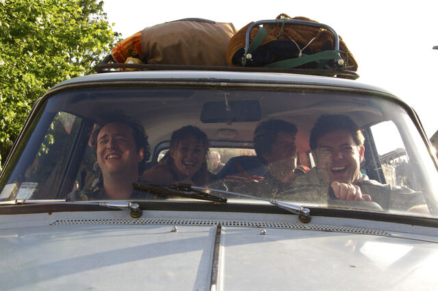 Vier mensen zitten in een auto. Ze lachen. Op het dak van de auto is bagage gebonden.