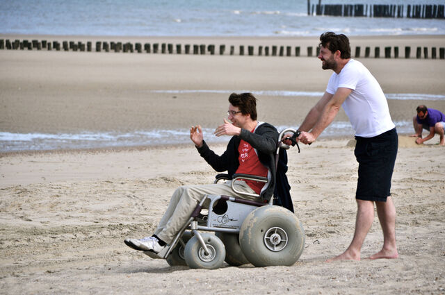 Een man zit in een rolstoel met enorm dikke banden. Hij bevindt zich o phet strand en wordt voortgeduwd door iemand op blote voeten, met een zwarte korte broek en een witte T-shirt