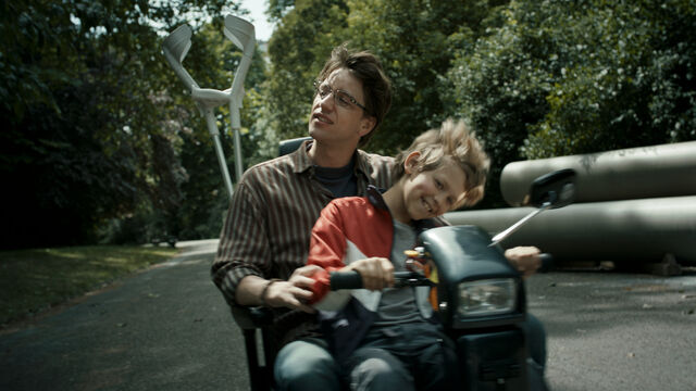 Een jongen zit op een elektrische rolstoel. Op zijn schoot zit een lachend kind. De wind waait door hun haren. Achterop de scooter zie je twee krukken.