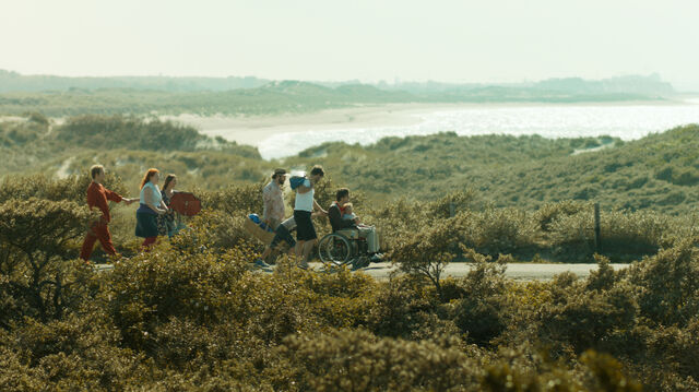 Foto van een groep mensen die in de duinen wandelen. Een iemand zit in een rolstoel die wordt voortgeduwd