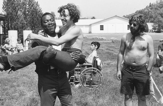 A bare-chested young man is carried by his friend. They are laughing.