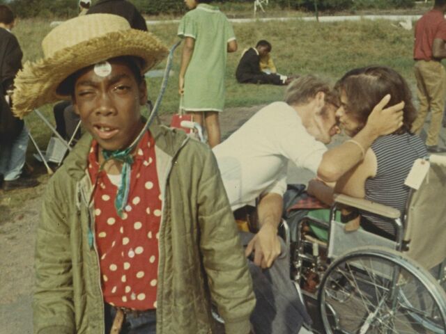 Een jongeman met een strohoed kijkt uitdagend in de camera. Op de achtergrond buigt een man zich over een vrouw in een rolstoel om haar een kus te geven.