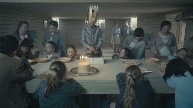 A little boy wearing a cardboard crown that has slipped over his eyes is opening his birthday present. His friends are sitting or standing around the table.