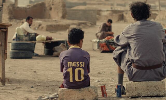 Hamoudi is sitting outside with his dad. He is wearing his Messi soccer jersey.