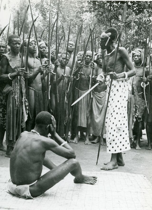 Een groep schaars geklede Congolezen staat rond een man die met zijn handen zijn hoofd vasthoudt