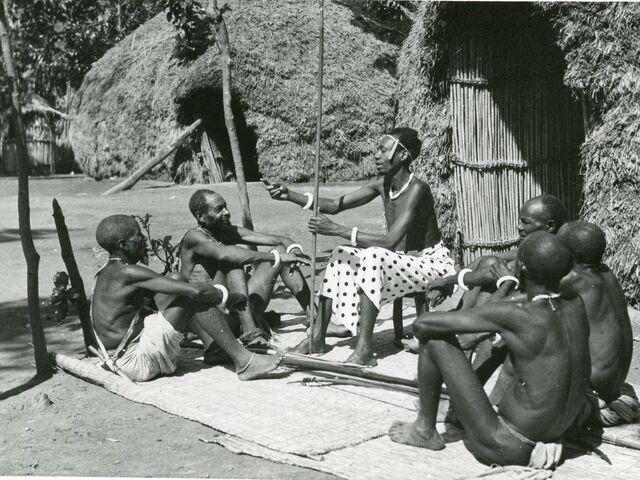 Een 5-tal Congolezen zit aandachtig te luisteren naar een zwarte man met een witte rok die een stok vasthoudt en op een stoel zit.