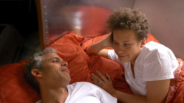 Jennifer en haar verloofde Omar liggen samen in bed. Jennifer steunt op haar elleboog, ze lachen naar elkaar. Ze dragen allebei een wit T-shirt.