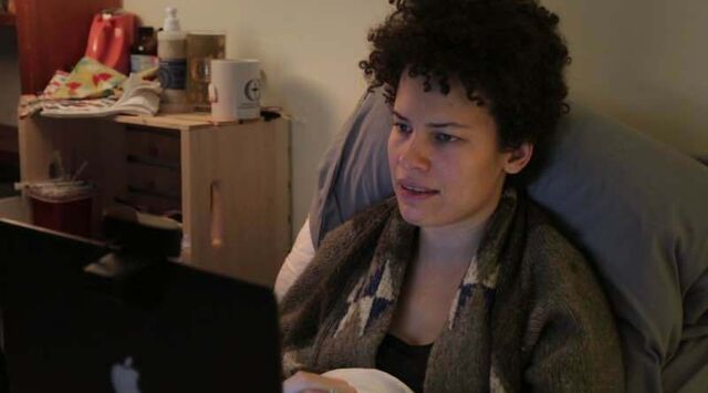 Jennifer zit in bed met haar laptop op haar schoot. Ze heeft donkere krullen en draagt een bruine trui.