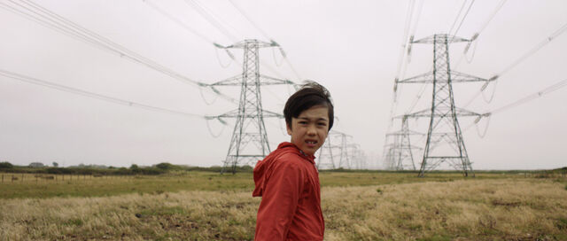 Een donkerharige jongen in een rode trui staat in een veld vol elektriciteitsmasten. Hij kijkt bezorgd.
