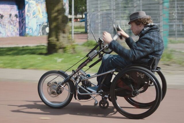Still uit de film 80% ongeschikt. Mari Sanders, een jongeman met bruin krulhaar, is op weg in zijn rolstoel waaraan een motorisch wiel is verbonden
