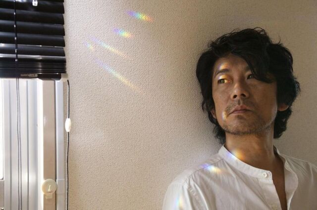 Still uit de film Radiance. Een man leunt tegen een witte muur. De lichtinval zorgt voor regenboogvlekjes op zijn gezicht en ogen.